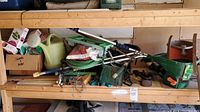 Shelf with various garden tools including Scotts seed spreader, watering can, hose, metal tools and seed box.