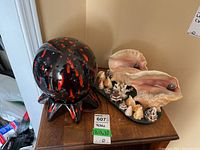 View of both lamps on wooden table: one black and red splattered ceramic lamp with round perforated shade, and one detailed shell-based lamp with conchs and many smaller shells on base.