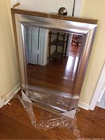 Full view of rectangular metal framed mirror with protective plastic on bottom corner, placed against a wall on hardwood floor.