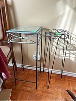 Photo showing two metal plant stands side by side. Left stand has glass top, rectangular shape with decorative metal sides. Right stand is round metal frame without glass.