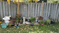 Wide outdoor shot showing multiple ceramic planters arranged along a wood fence with plants and soil included, plus a white birdbath on the left