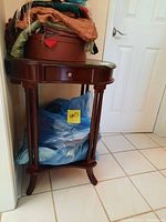 Side table shown against a wall with various cloth and hat items on top and plastic bags below. Photo shows drawer knob, bottom shelf, and leg design.
