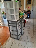Four plastic storage units, two gray with translucent drawers and two white units stacked above, placed in a tiled hallway area with some clutter in the background.