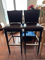 2 black bar stools with cushioned seats, nailhead trim, and wooden legs with footrests seen in front of window.