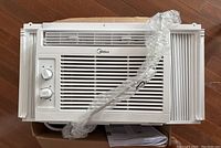Front view of white Midea window air conditioner showing mechanical dial controls and front air grille with side window fitting panels and wrapped flexible exhaust hose.