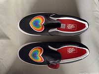 Top-down view of black Vans slip-on sneakers with rainbow heart embroidery on each shoe