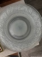 Top view of a clear glass dinner plate showing the intricate floral and geometric textured pattern on the plate's edge and center.