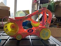 Side view of a large colorful Littlest Pet Shop plastic pet vehicle, coral with floral designs, yellow wheels, and a clear dome top, featuring a yellow rotating drum wheel on one side and a hanging swing inside.