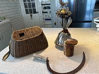 Four antique items on a countertop: metal garden art flower, marble jar with lid, rusty sickle with bone handle, and woven creel basket with fabric strap.