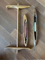 Photo of antique niddy noddy spinner and two wooden weaving shuttle boats with thread spools laid on wooden floor.