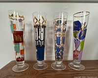Four tall Ritzenhoff beer glasses arranged on wood surface showing unique colorful designs and gold accents.