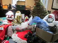 Wide view showing tall green Christmas tree, two Santa figurines, wrapped gifts, wreath, and Rubbermaid tote filled with decorations and lights.
