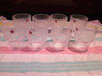 Set of eight clear textured Iittala Paadar shot glasses arranged in two rows on a pink striped towel, each with red Iittala stickers visible.