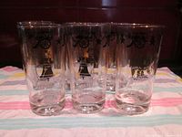 Frontal view of six clear tall glasses each decorated with black and gold Americana-themed decals, showing the Liberty Bell, hats, wagon, and stars.