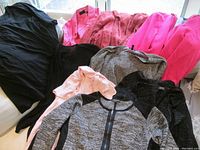 Nine women's clothing items displayed on surface, mixture of jackets, sweaters, and shirts in various colors including black, pink, beige, and gray.
