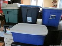 Photo showing six large storage totes of various colors (green, blue, dark blue) arranged on a table; four have lids and two do not. One white lid is visibly cracked.