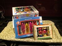 Five boxes of vintage Christmas ornaments stacked, showing original packaging with colorful graphics and text such as Santa Land and Christmas tree ornaments.