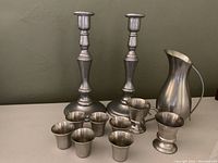 Group photo showing two tall pewter candlesticks, six small shot glasses, a creamer, sugar bowl, and a tall pitcher on a table against a green wall.