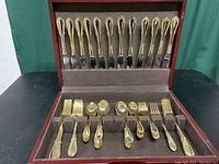 Open wooden case displaying gold electroplated flatware pieces organized neatly in slots with 12 table knives in upper section and forks, spoons in lower section.