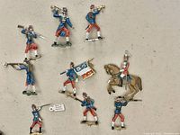 Close-up view of 11 metal miniature soldier figures including two with flags, one mounted on horse; painted details in blue, red, white; some with rifle, horn, or sword.