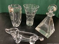 Photo showing all four crystal pieces together on black surface with green background: Kosta Boda vase, Waterford vase, Dachshund candy dish, and Vannes le Chatel decanter with stopper.