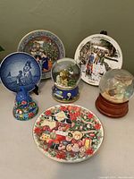 Wide view of all seven items: four collector plates of Christmas themes, two snow globes each musical, and a hand-painted pottery bell.
