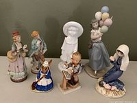 Seven vintage porcelain figurines displayed on a flat surface including a Lladro Balloon Girl, a white glazed Spode girl with teddy bear, an Occupied Japan couple, a Hummel conductor, a Goebel Madonna and Royal Doulton Mrs Bunnykins.