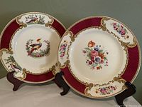 Two Myott dinner plates displayed on stands showing the Chelsea Bird plate and Bouquet floral plate with red and gold border design.