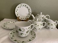 Full view of the seven piece tea set including teapot with lid, sugar bowl with lid, creamer, cup with saucer, and two bread and butter plates. All items decorated in green clover pattern on white porcelain.