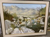 Front view of the large oil painting depicting a stream bed with mountains and natural foliage with beige frame and matting.