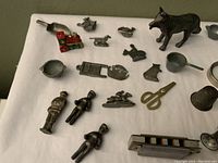 Multiple antique metal miniatures on fabric background showing dog figurine, harmonica, steam engine, magnifying glass, scissors, thimble, ladle, and various small figures and items.