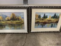 Pair of framed pastel landscape paintings showing scenes with trees, water reflections, and mountains under glass in silver frames.