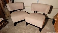 Full view of two beige upholstered accent chairs with curved backrests and wooden legs placed on carpeted floor.