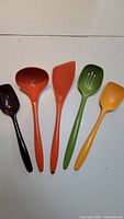 Five colorful plastic cooking utensils laid out side by side on white surface