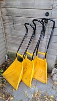 Three yellow Garant snow shovels with black handles standing against a wooden wall on pavement with leaves scattered on the ground.
