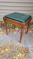 Side view of vintage wooden piano stool with green padded cushion, shown outdoors with fallen leaves.