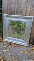 Frontal photo of a square silver framed mirror reflecting outdoor greenery and fall foliage, showing overall size and frame style.
