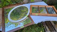 Three mirrors arranged on a white cloth outdoors, showing the oval silver framed mirror, one large rectangular wooden framed mirror, and one rectangular old window style mirror with three glass panes.