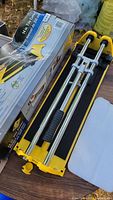 Photo showing the tile cutter laid out with close view of the metal rails, cutting handle, yellow plastic ends, and original packaging box next to it.