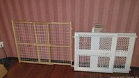 Wooden mesh baby gate and white plastic gate stacked side by side against a wall with wood floor and patterned wallpaper.