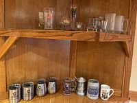 Shelf with assorted mugs including metal tumblers, decorative beer steins, and ceramic mugs.