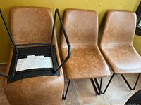 Front view of three light brown faux leather dining chairs and one chair overturned showing black metal frame and underside construction.