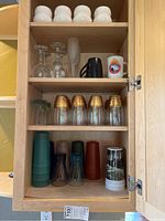 Glassware collection including white bowls, wine glasses, mugs, and glass tumblers with gold rim on wooden shelves inside kitchen cabinet.