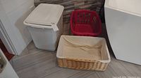 Photo of a white plastic laundry hamper with lid, rectangular woven basket with liner, and small red plastic basket placed on a floor near a washing machine.