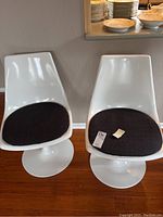 Two white tulip swivel chairs side-by-side with black cushions on a wood floor against a gray wall.