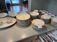 Photo showing the full collection of 87 Meito Japan china pieces arranged on a counter including stacked plates, bowls, cups, and serving pieces.