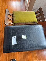 Top view showing one black crocodile-pattern ottoman bench with a green cushion resting on its wooden slatted backrest, and another bench next to it, on wooden floor.