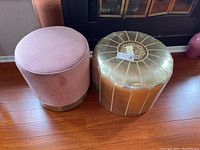 Two ottomans placed side by side on a wood floor; one pink velvet with gold base, the other gold leather with stitched segments and decorative embroidery.