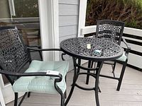 Two black metal bar stools with light seafoam green cushions and round metal patio table with lattice design top.