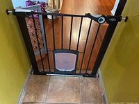 Full view of black metal pet/baby gate installed in doorway with pressure mounts and built-in pet door with paw print.